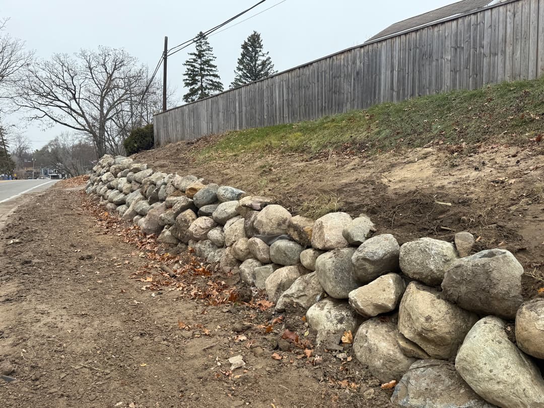 Stone retaining wall along a dirt path beside a road and wooden fence on a cloudy day.