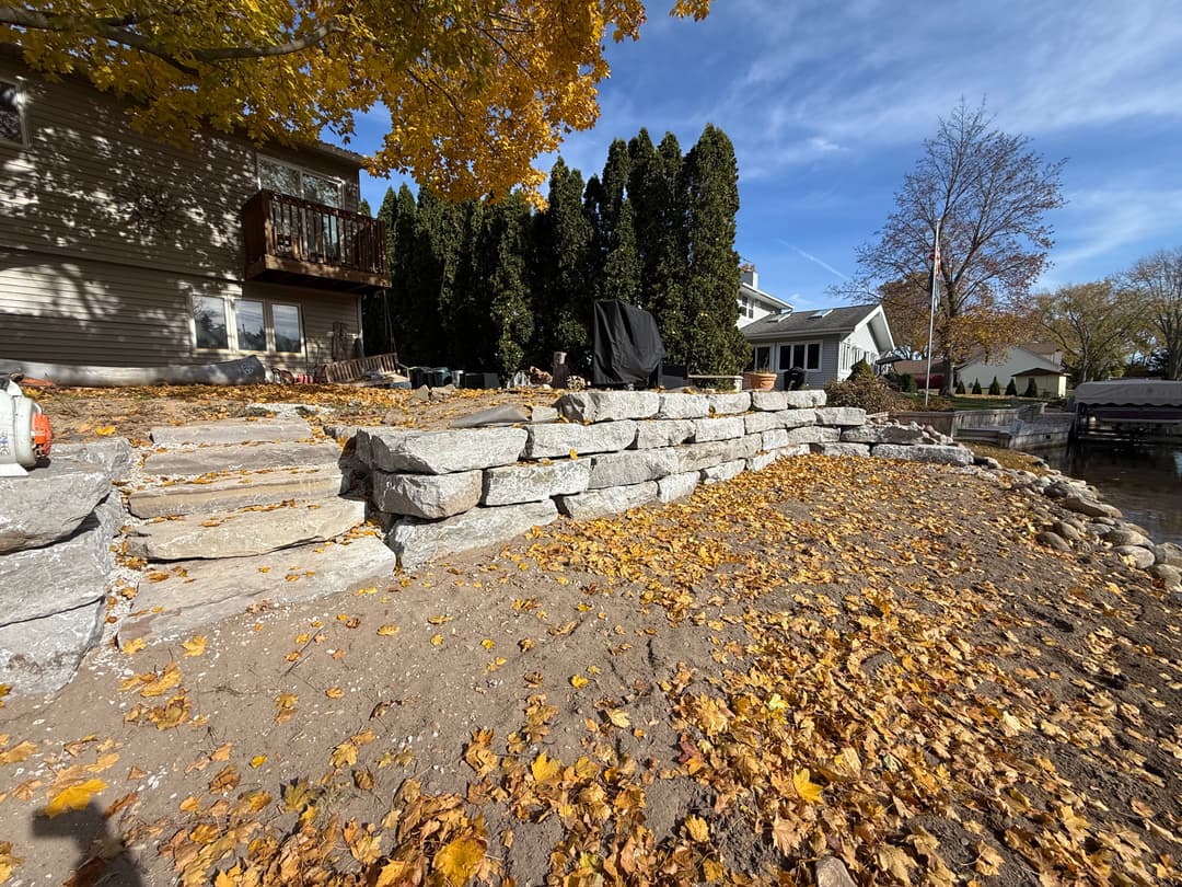 Stone retaining wall with autumn leaves, alongside a waterfront property and clear blue sky.