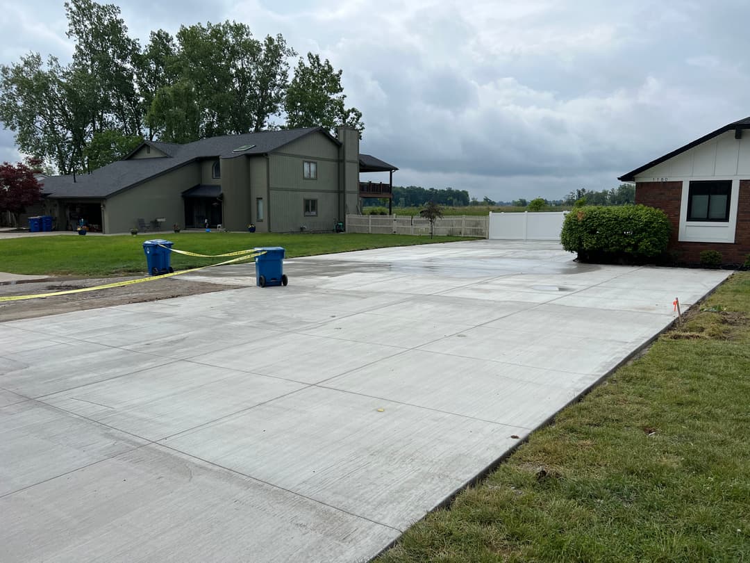 Newly poured concrete driveway with blue trash bins, grassy areas, and residential homes nearby.