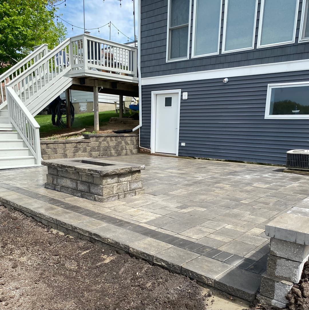 Paved patio area with fire pit next to a home featuring a gray exterior and wooden deck.
