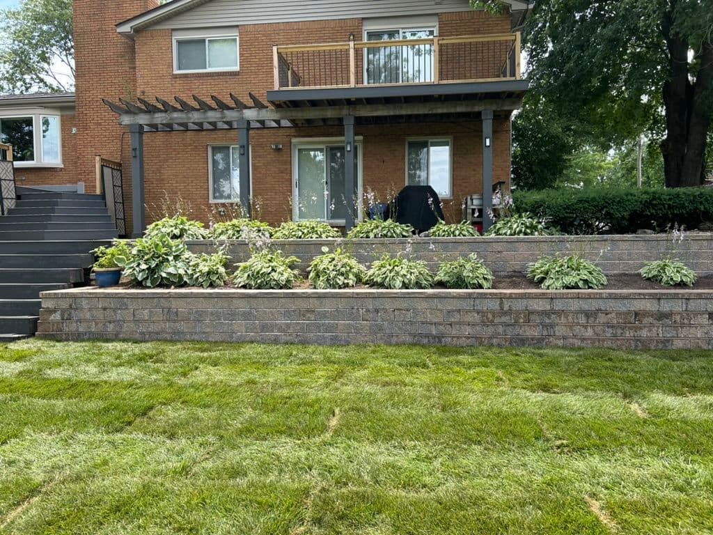 Backyard landscape featuring tiered stone garden beds, green shrubs, and a modern home facade.