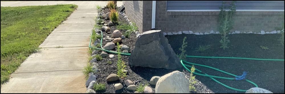 Landscape design featuring stones, plants, and a green garden hose along a sidewalk.