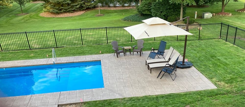 Backyard pool with lounge chairs and umbrella, surrounded by green lawn and fencing.