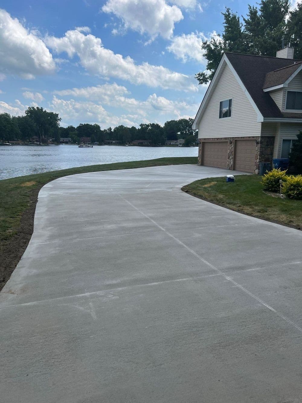 Newly poured concrete driveway beside a serene lake and residential house on a sunny day.