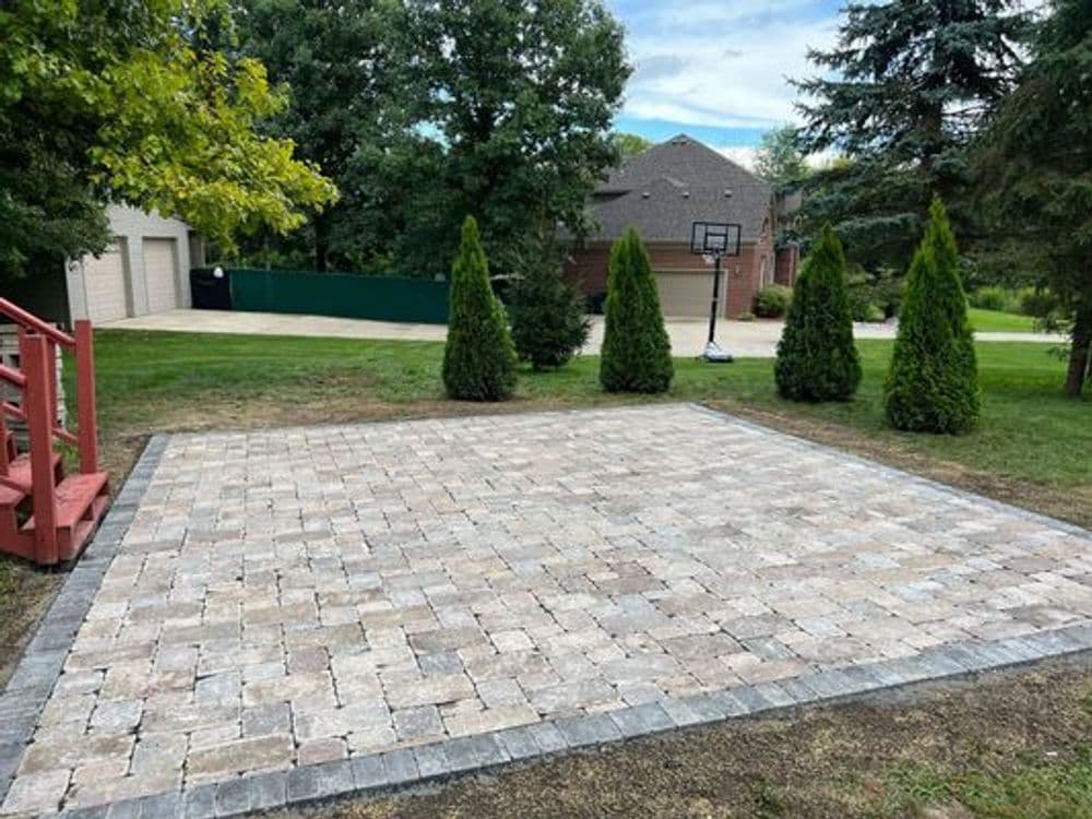 Paver patio installation in a backyard with green trees and a basketball hoop nearby.
