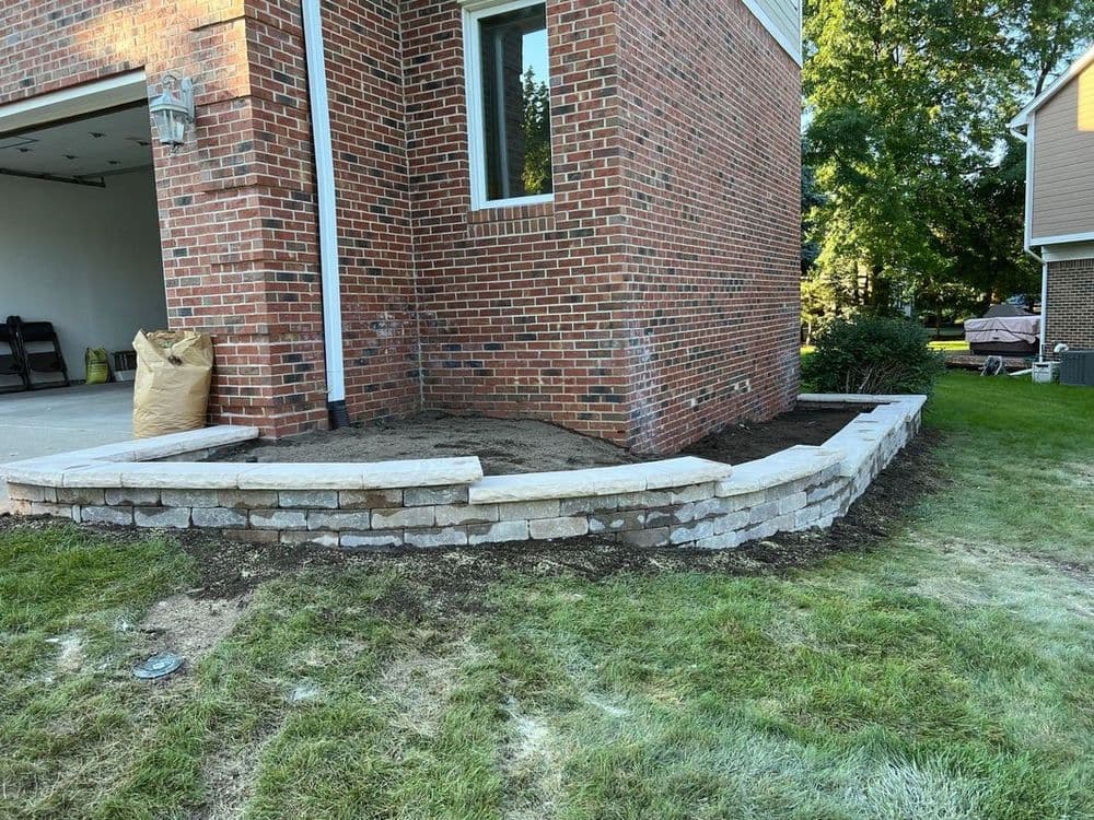 Stone retaining wall under construction beside a brick house with fresh soil and landscaping.