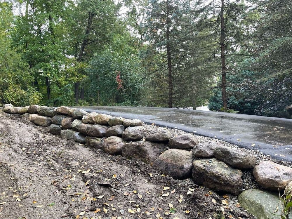 Stone retaining wall beside a gravel road lined with trees and foliage.