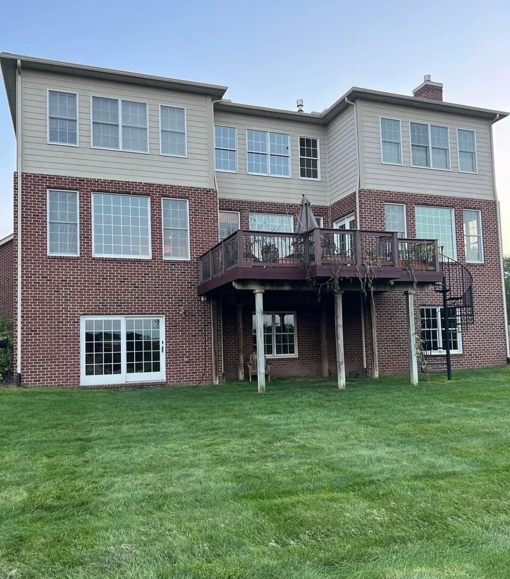Two-story brick house with large windows and spiral staircase leading to deck, surrounded by green lawn.